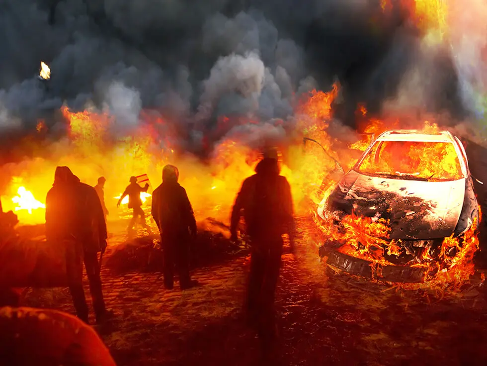 Individuals near a vehicle on fire with smoke in the background