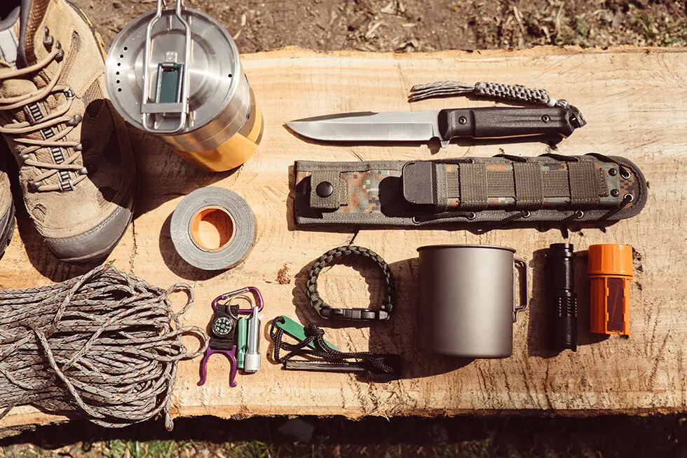Overhead top view of hiking travel gear on wood log. Items include hiking boots, cup, rope, knife, matches,
        flashlight, compass. Flat lay of outdoor travel equipment items for mountain camping trip