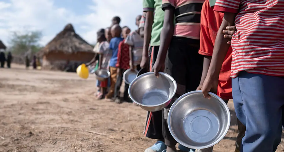 Queue of people, likely in a rural area, holding empty bowls, possibly waiting for food distribution