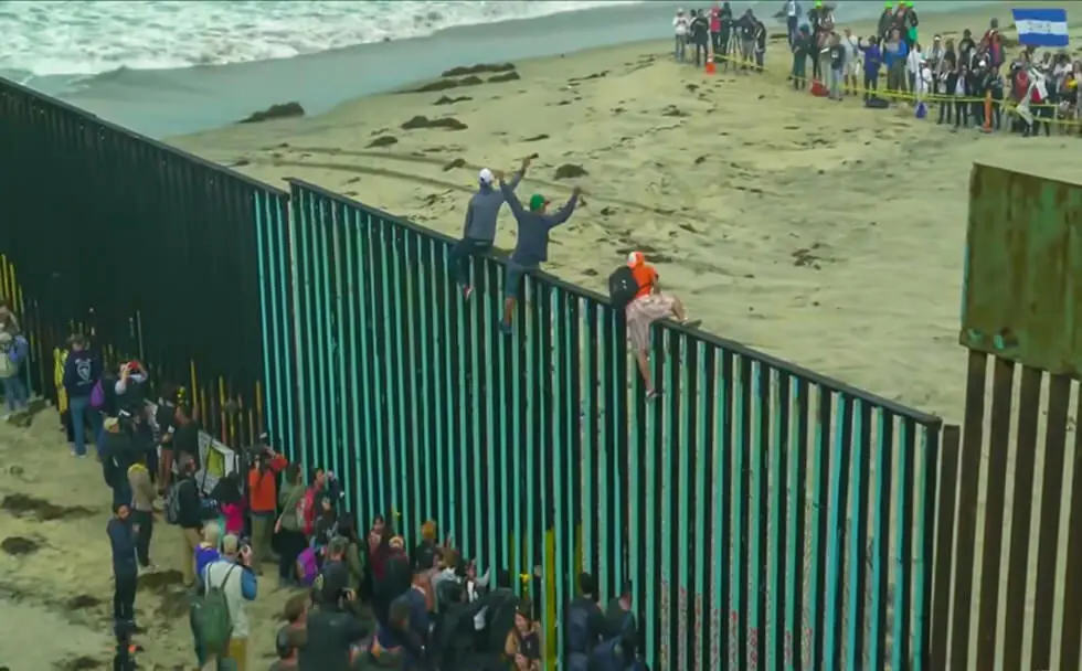 People gathered at a tall border fence by the beach, with some climbing on it